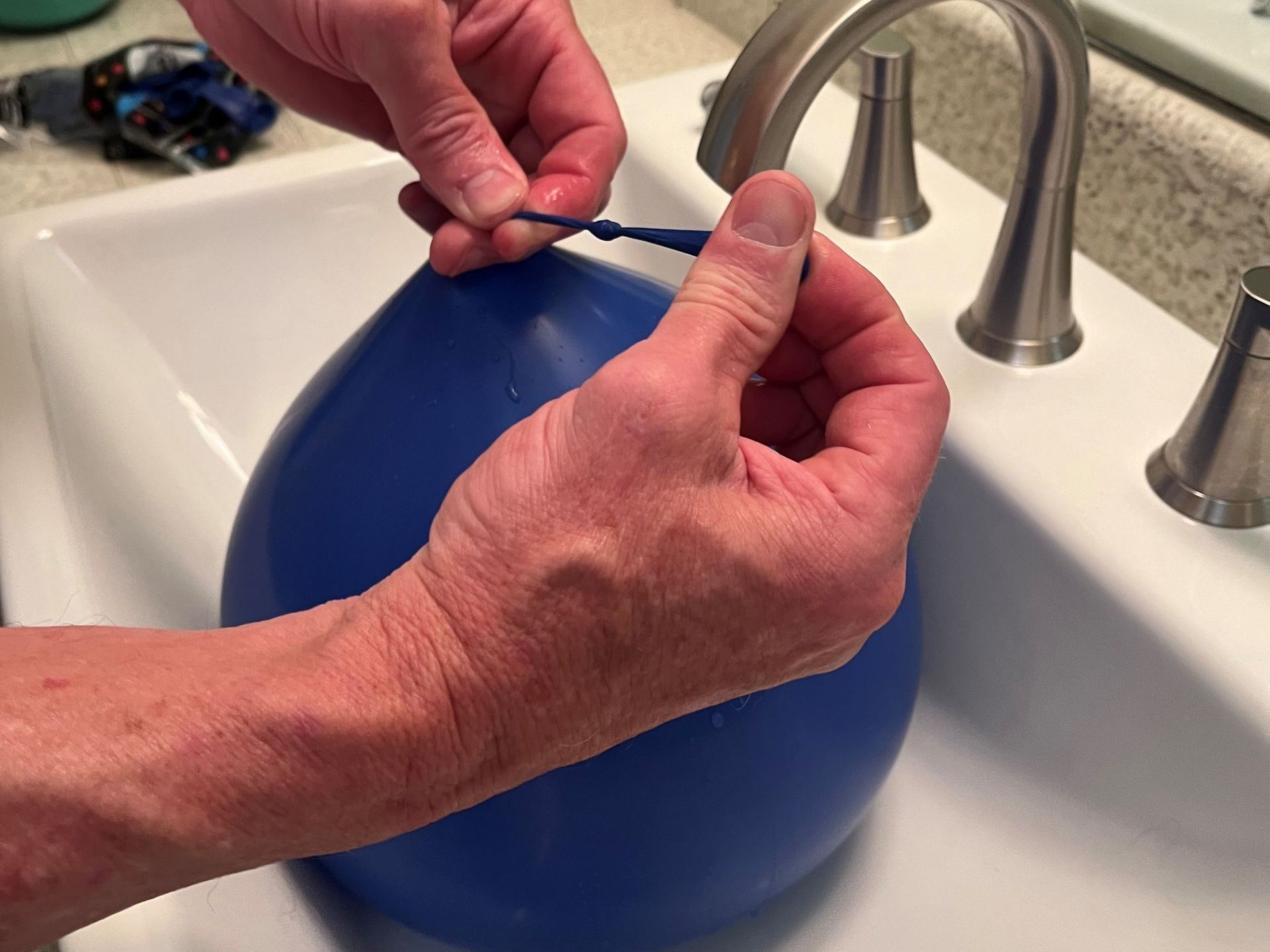 Hands tying a knot on a blue water balloon filled with water in a white bathroom sink.