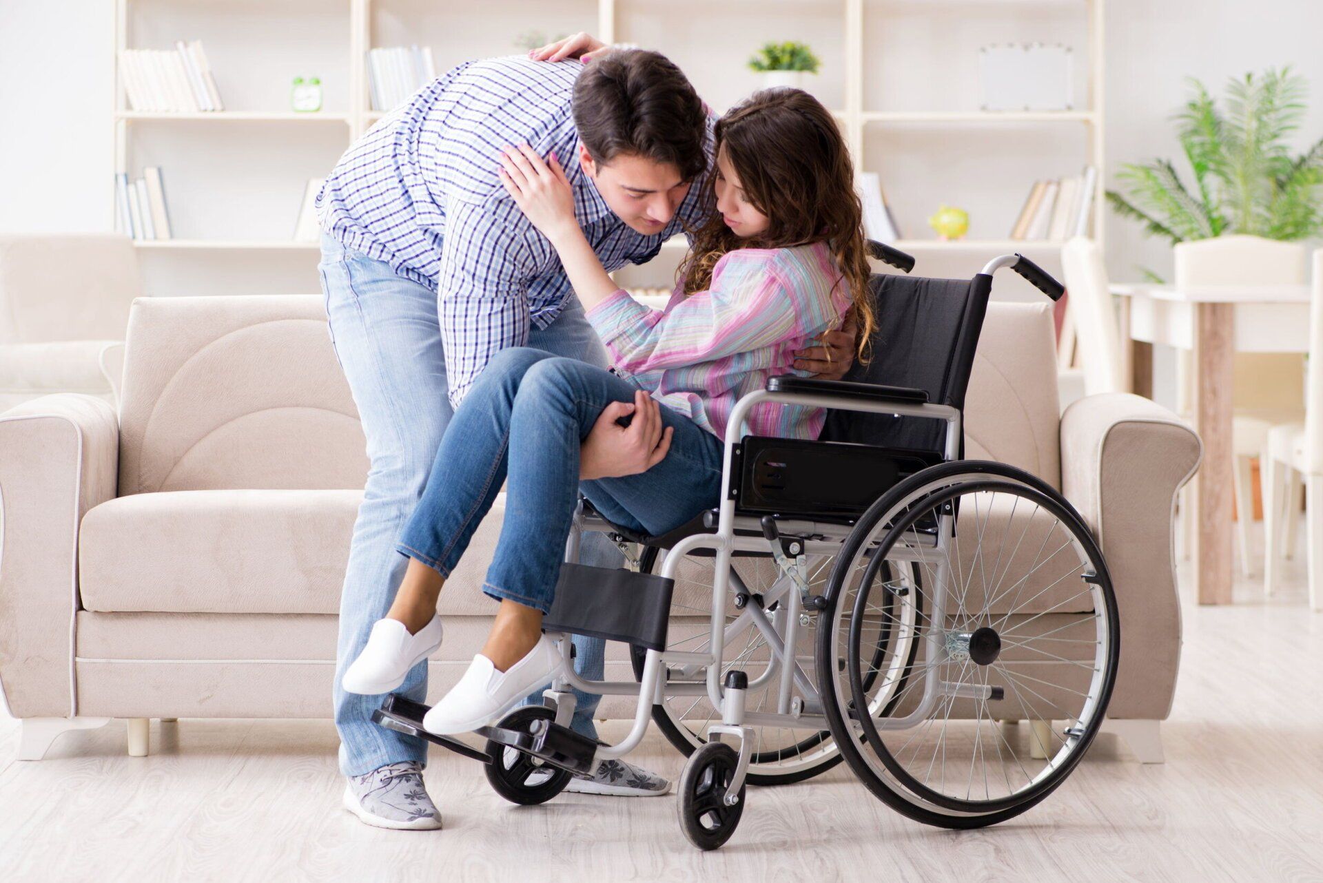 A man is carrying a woman in a wheelchair in a living room.