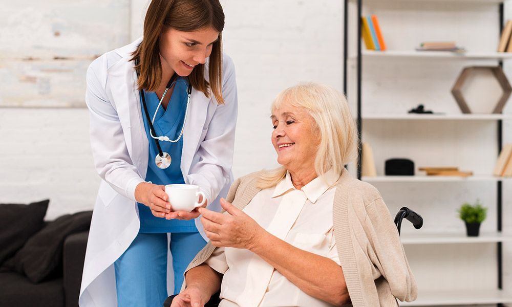A nurse is giving an elderly woman in a wheelchair a cup of coffee.