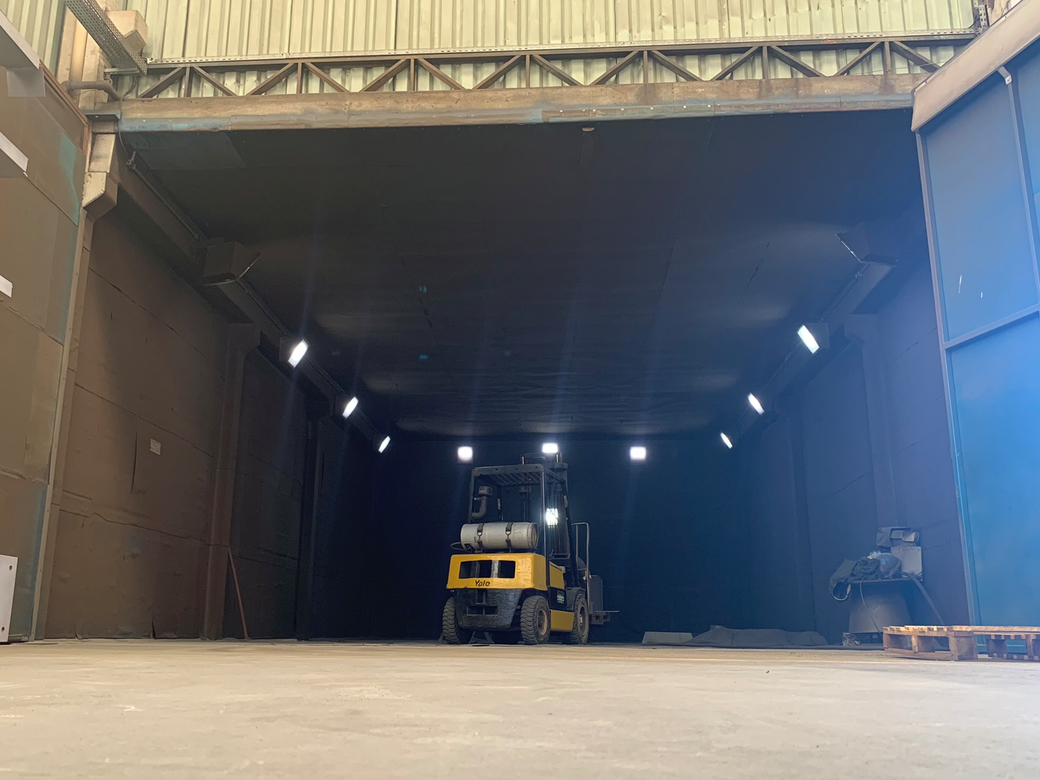 Carregadeira amarela dentro de um armazém mal iluminado com luzes no teto e uma entrada aberta.