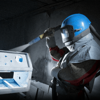 Operário com capacete azul usando uma ferramenta elétrica ao lado de uma máquina em um ambiente industrial pouco iluminado.