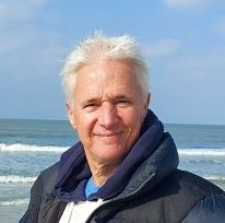 Een glimlachend persoon in een donkere jas staat aan de oceaan onder een blauwe hemel.