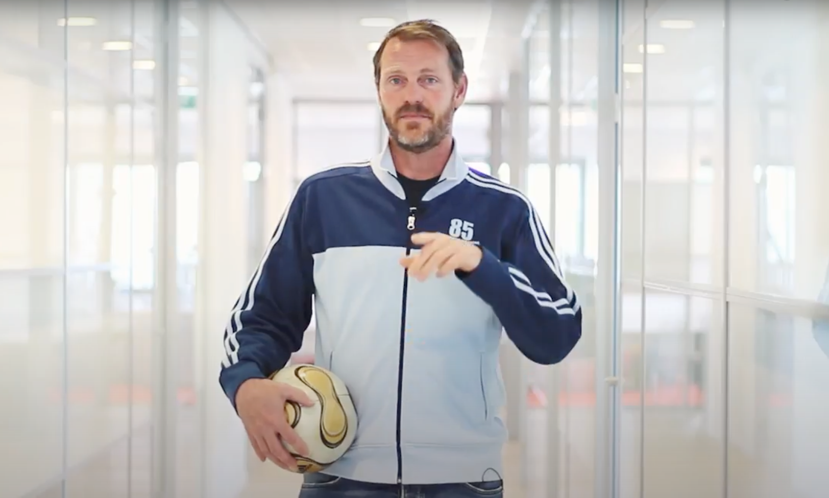Een man in een blauw trainingsjack houdt een voetbal vast in een lichte gang.