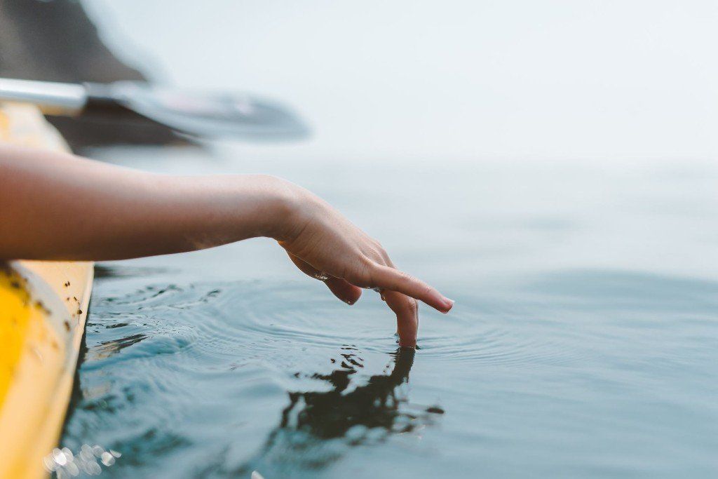 Een hand raakt het water aan vanuit een gele kajak, met een peddel op de achtergrond. Kalm water, mistige atmosfeer.
