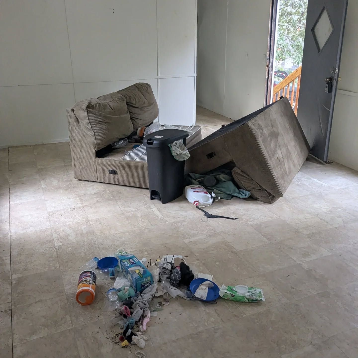Room with debris: upturned couch, overflowing trash can, scattered trash on floor, open door to outdoors.