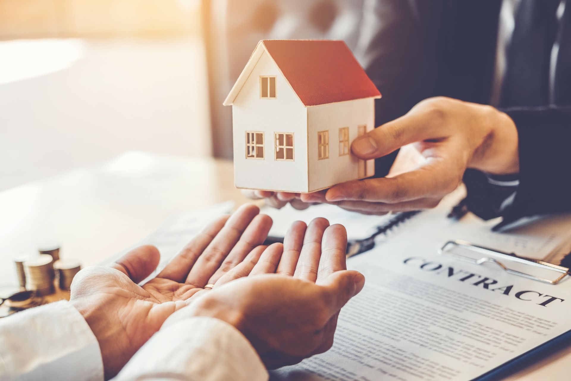 Person handing a miniature house to another over a contract, representing a real estate transaction.
