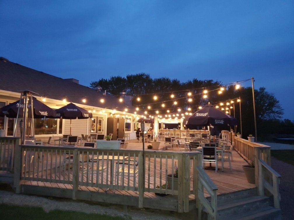 An outdoor patio deck at dusk, illuminated by strings of warm lights above tables, chairs, and large umbrellas.