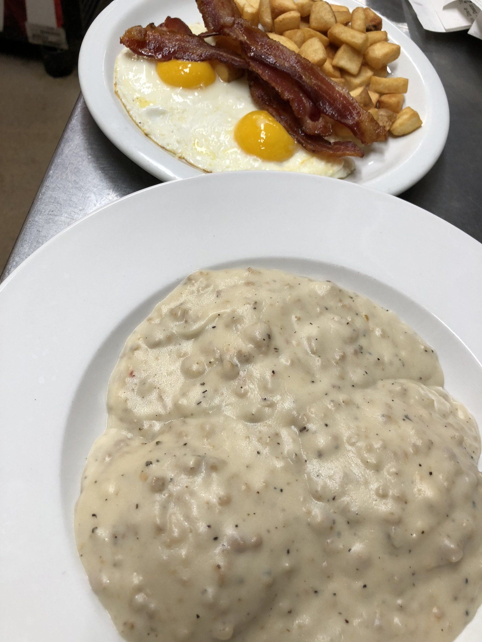 A close-up of a breakfast meal: a bowl of biscuits and gravy, with a plate behind containing eggs, bacon, and potatoes.