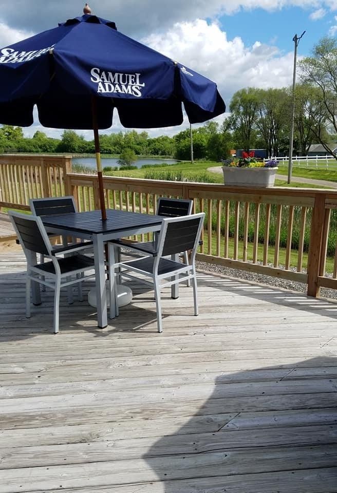 A square patio table and four chairs under a blue Samuel Adams umbrella on a wooden deck overlooking a pond.