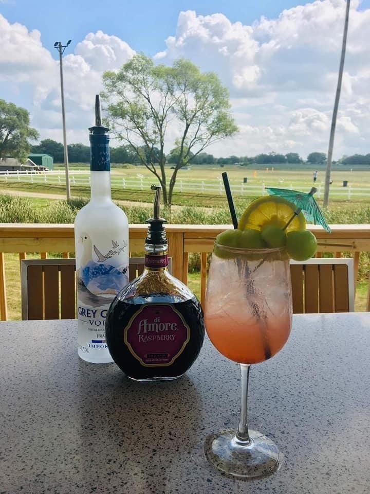 A cocktail, a bottle of vodka, and a bottle of liqueur sit on a table outdoors with a scenic field and tree background.