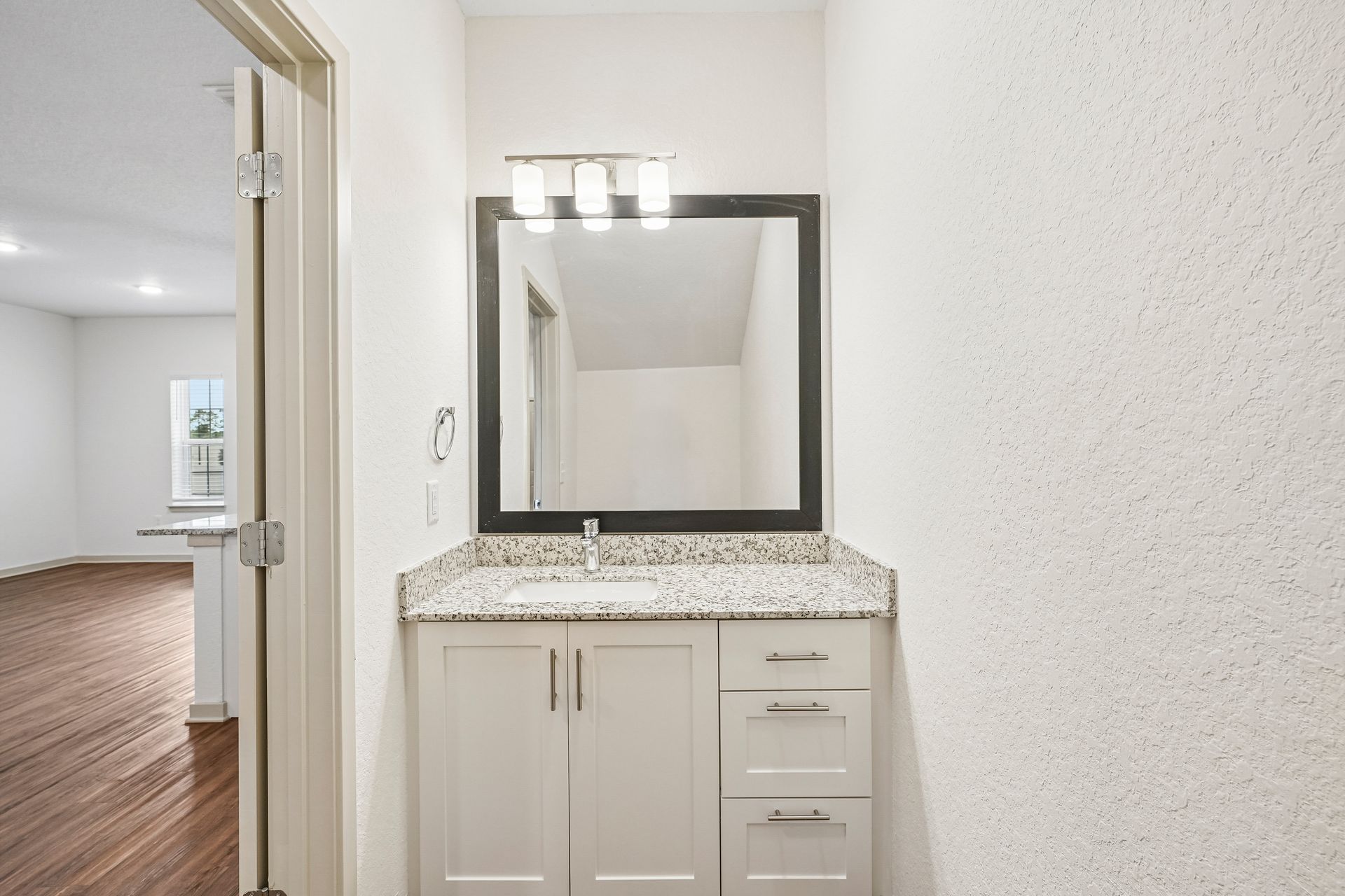 Bathroom vanity with white cabinets, speckled countertop, and black-framed mirror near an open doorway