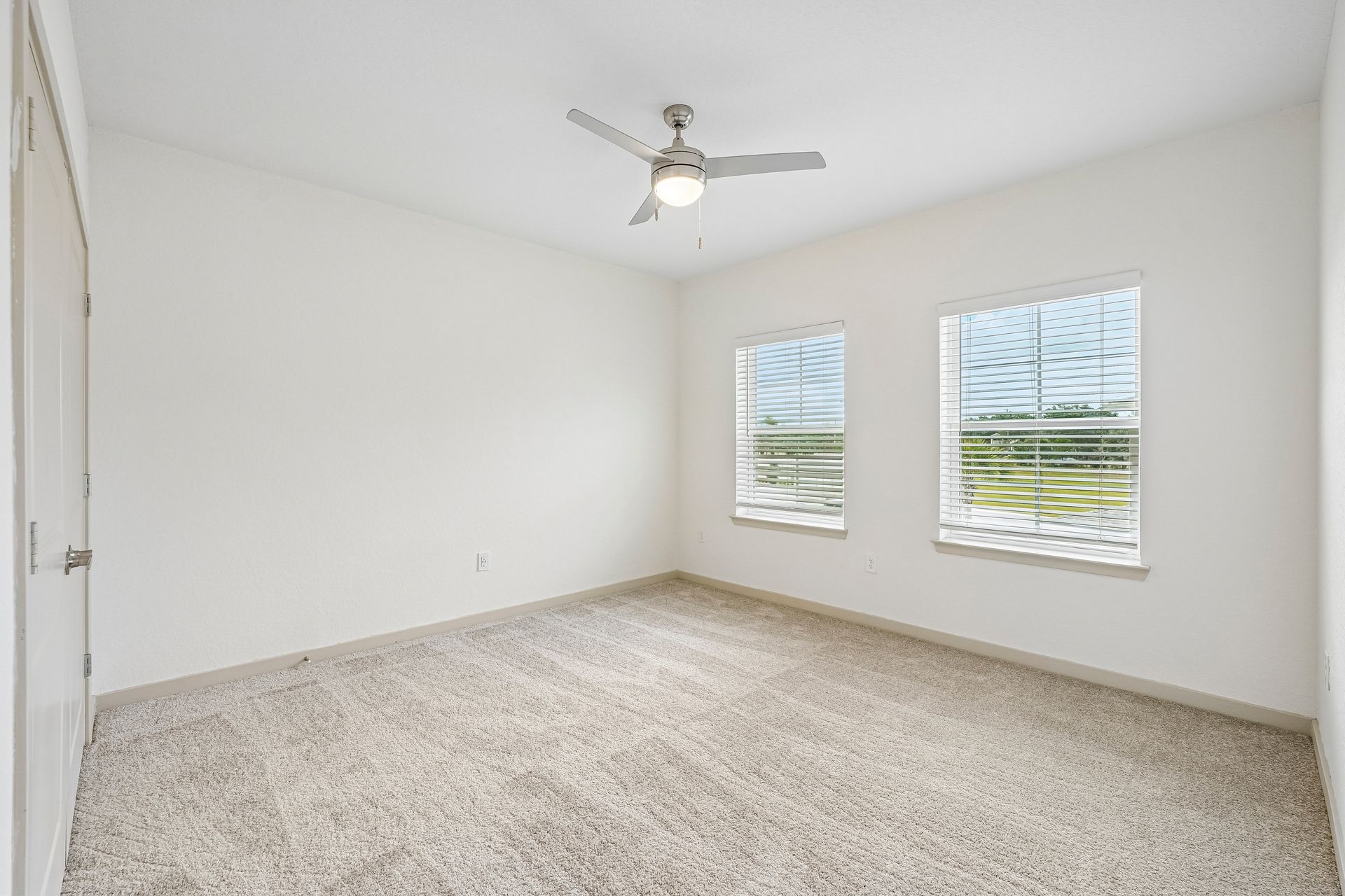 Empty white bedroom with carpet, ceiling fan, and two windows with blinds