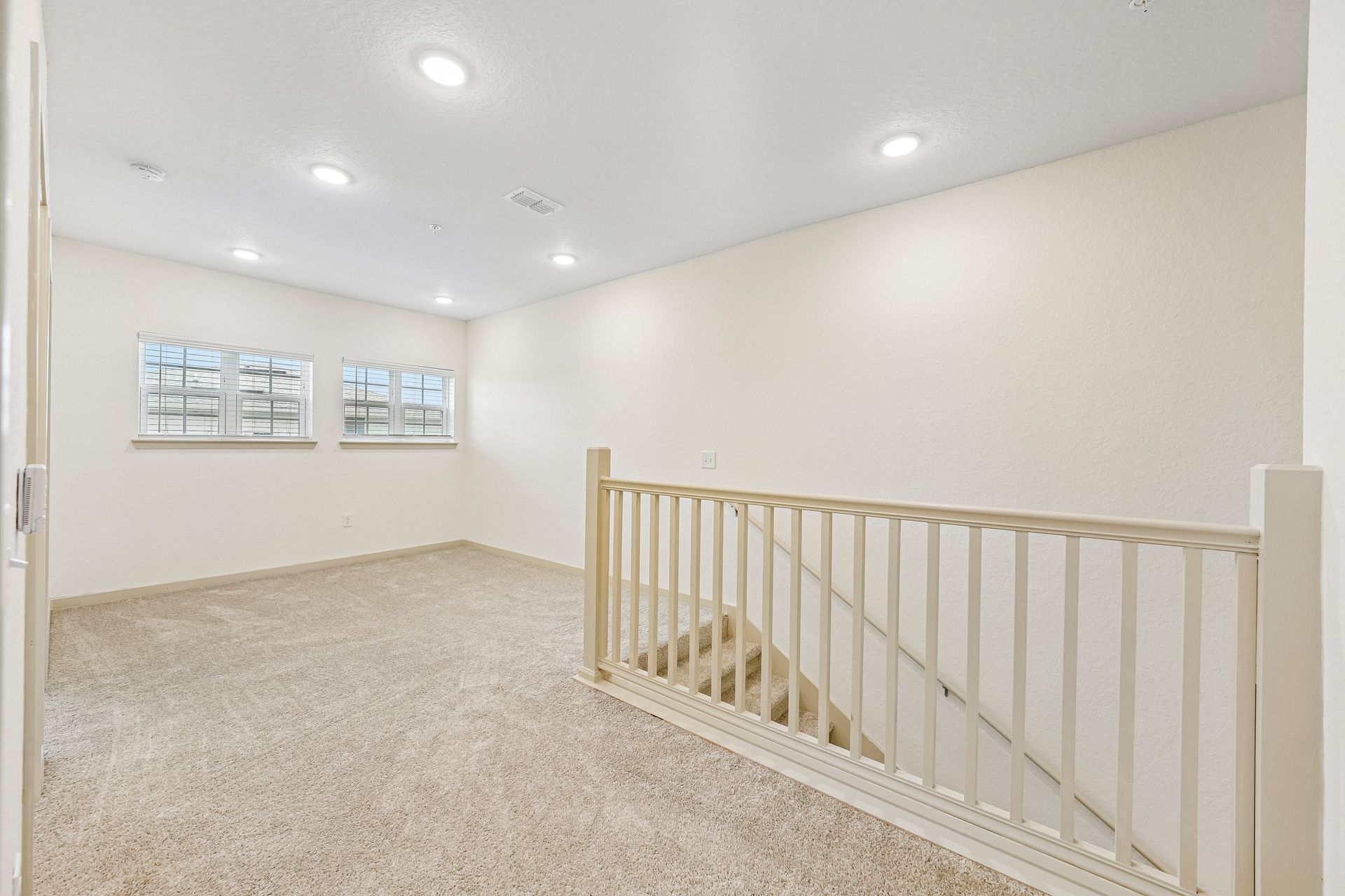 Bright carpeted upstairs landing with white walls, recessed lights, and stair railing beside two small windows