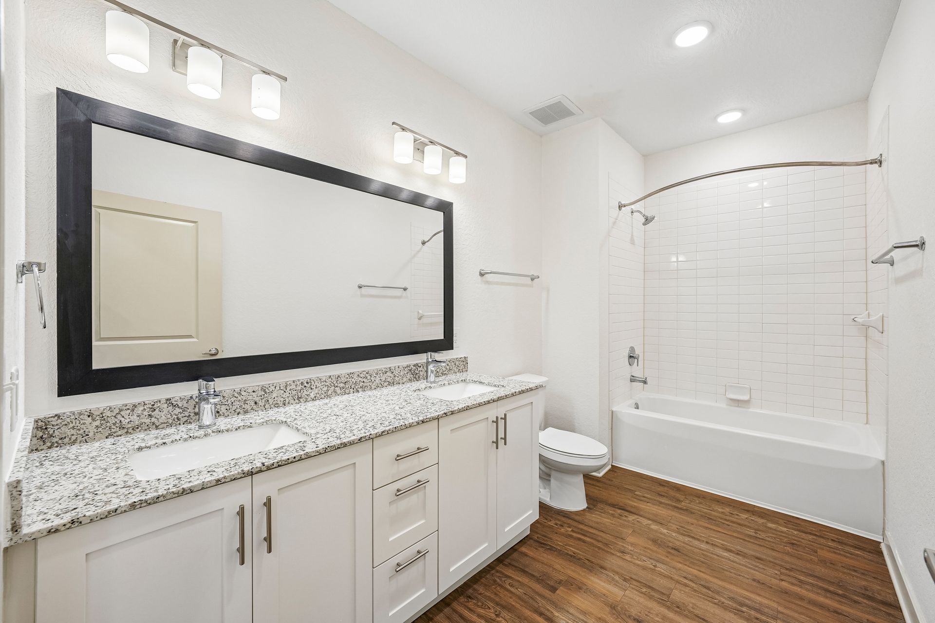 Modern bathroom with double vanity, large mirror, white tub, and wood-look floor.