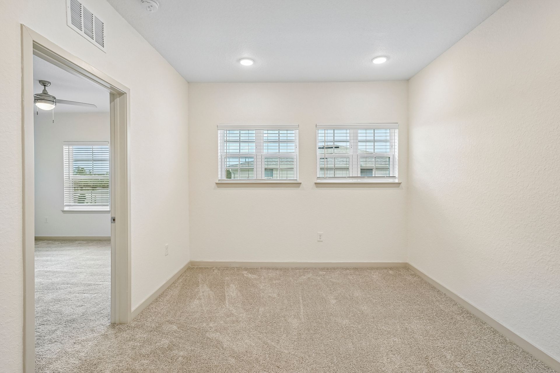 Empty beige room with carpet, two small windows, and an open doorway to a bright adjacent room