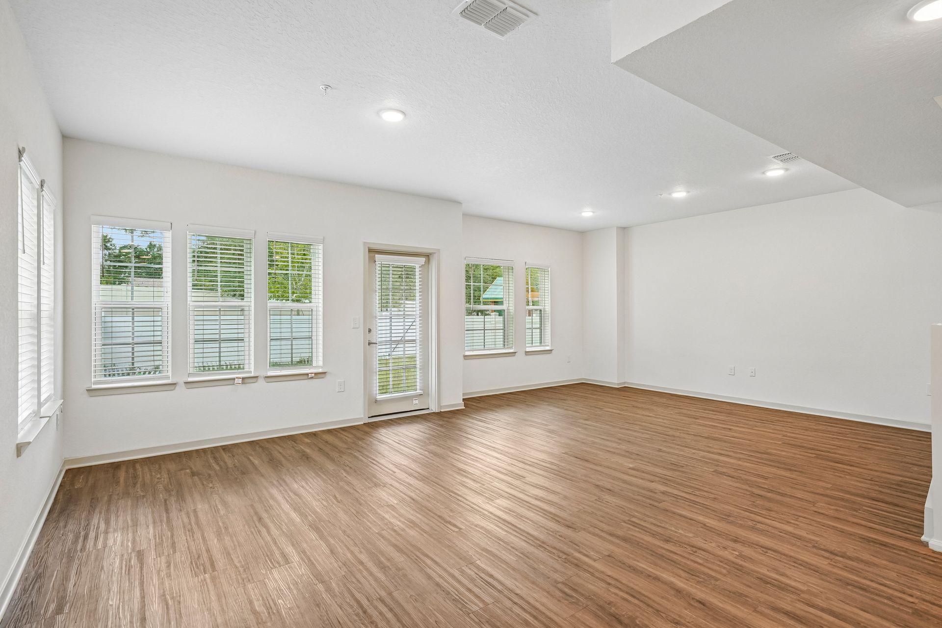Empty bright living room with wood floors, white walls, recessed lights, and windows with a glass door.