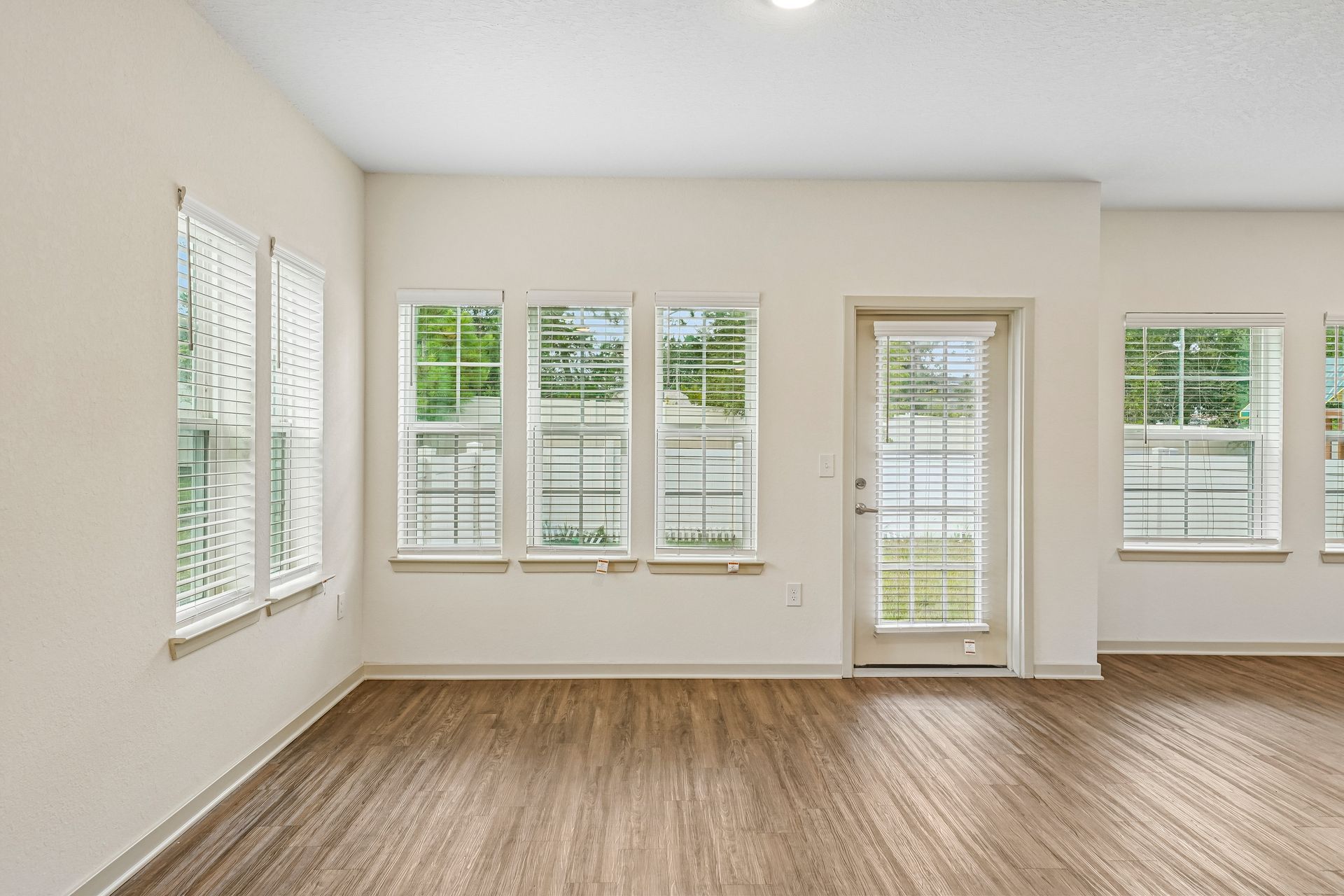Bright empty room with beige walls, wood flooring, and multiple white-trimmed windows and glass door.