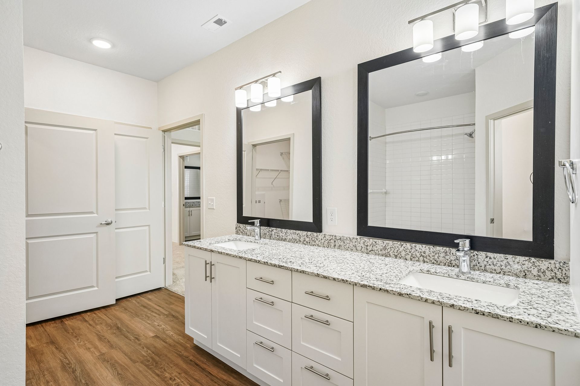 Bright bathroom with double vanity, large mirrors, white cabinets, and wood-look tile floor