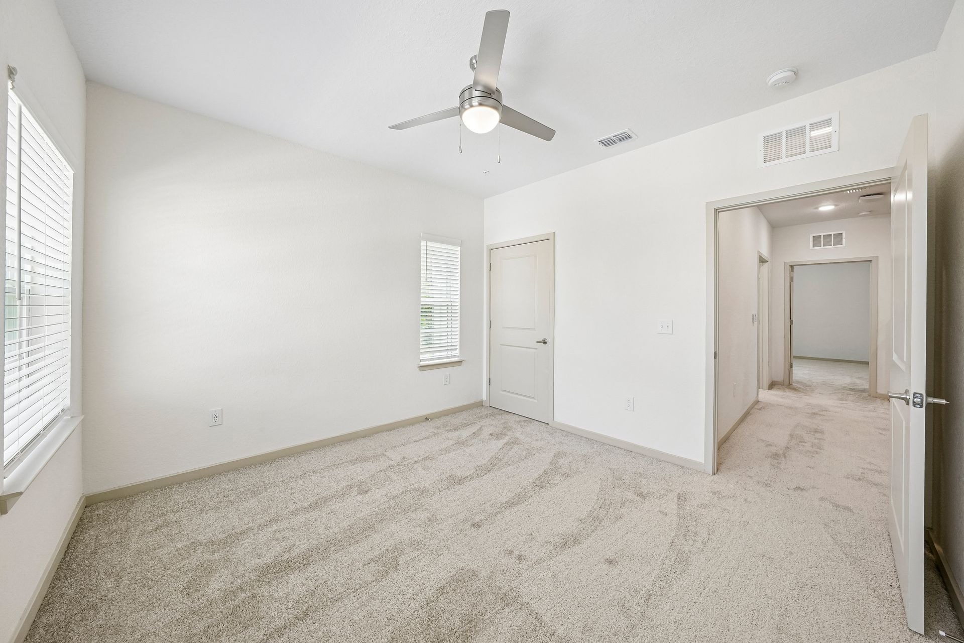 Empty beige carpeted bedroom with white walls, ceiling fan, and open doorway to a hallway