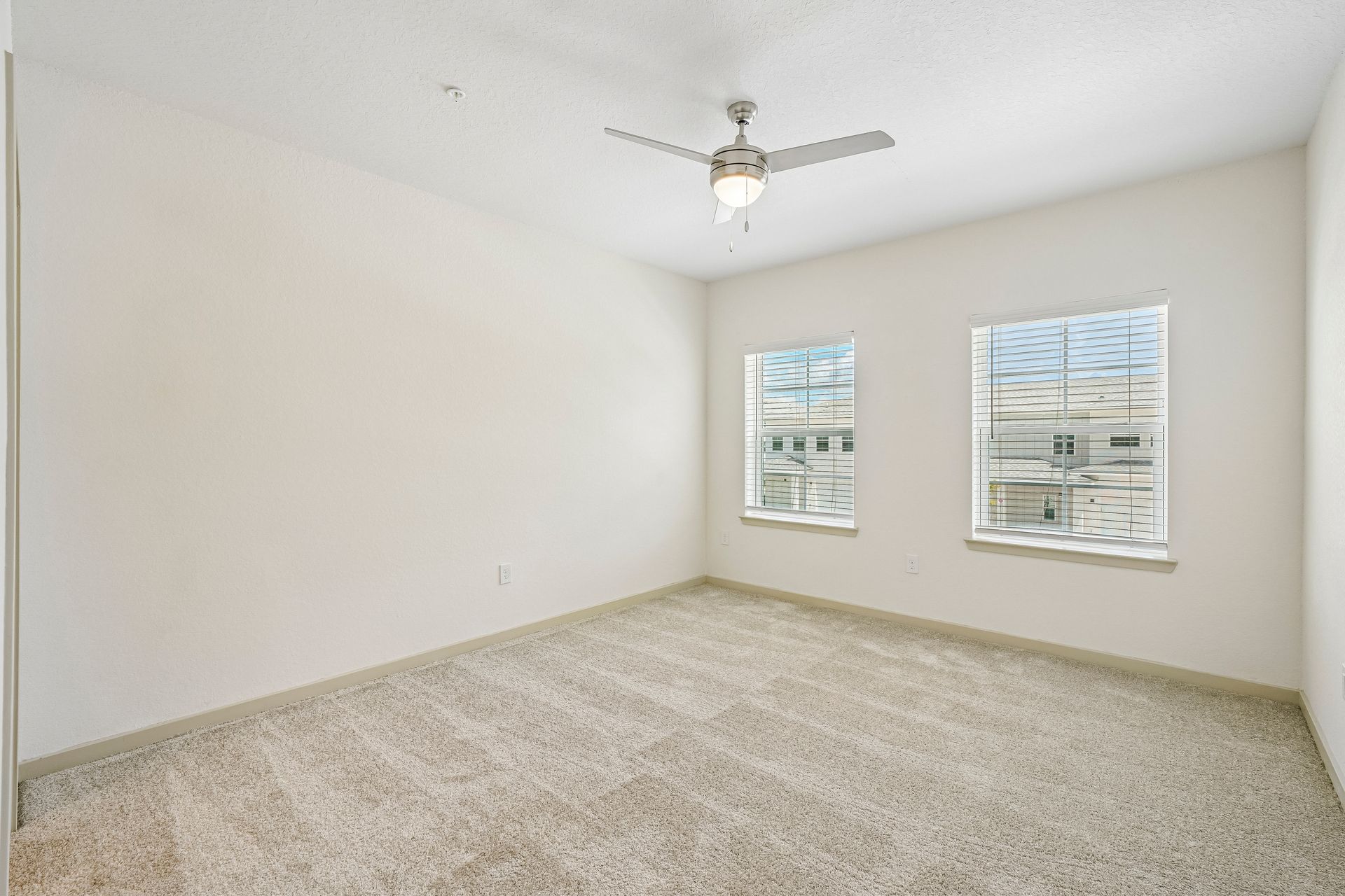 Empty beige bedroom with carpet, white walls, two windows, and a ceiling fan