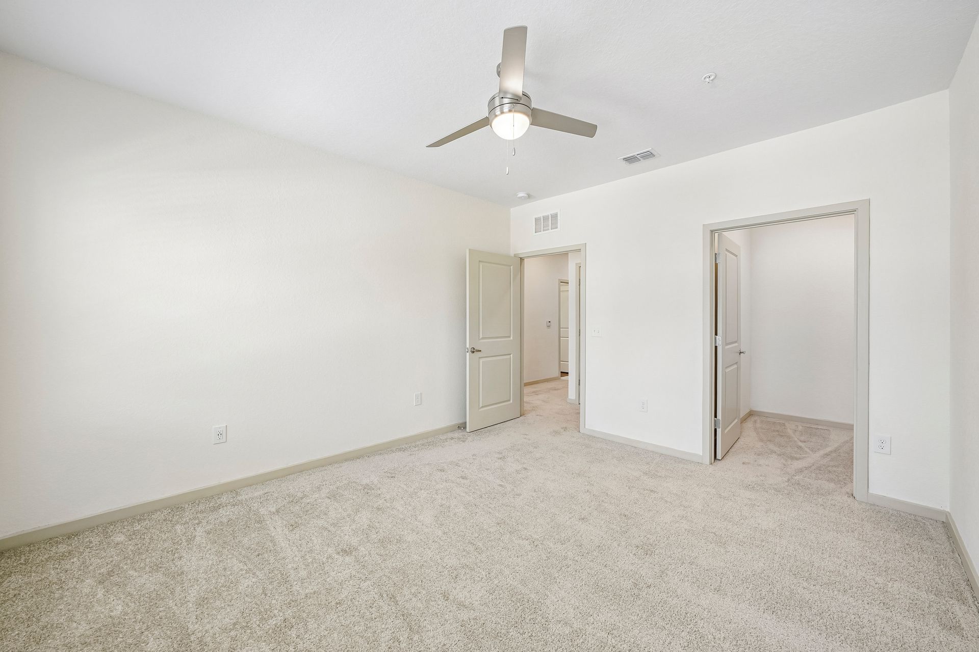 Empty white bedroom with carpet, ceiling fan, and two open doorways to a bathroom and closet area
