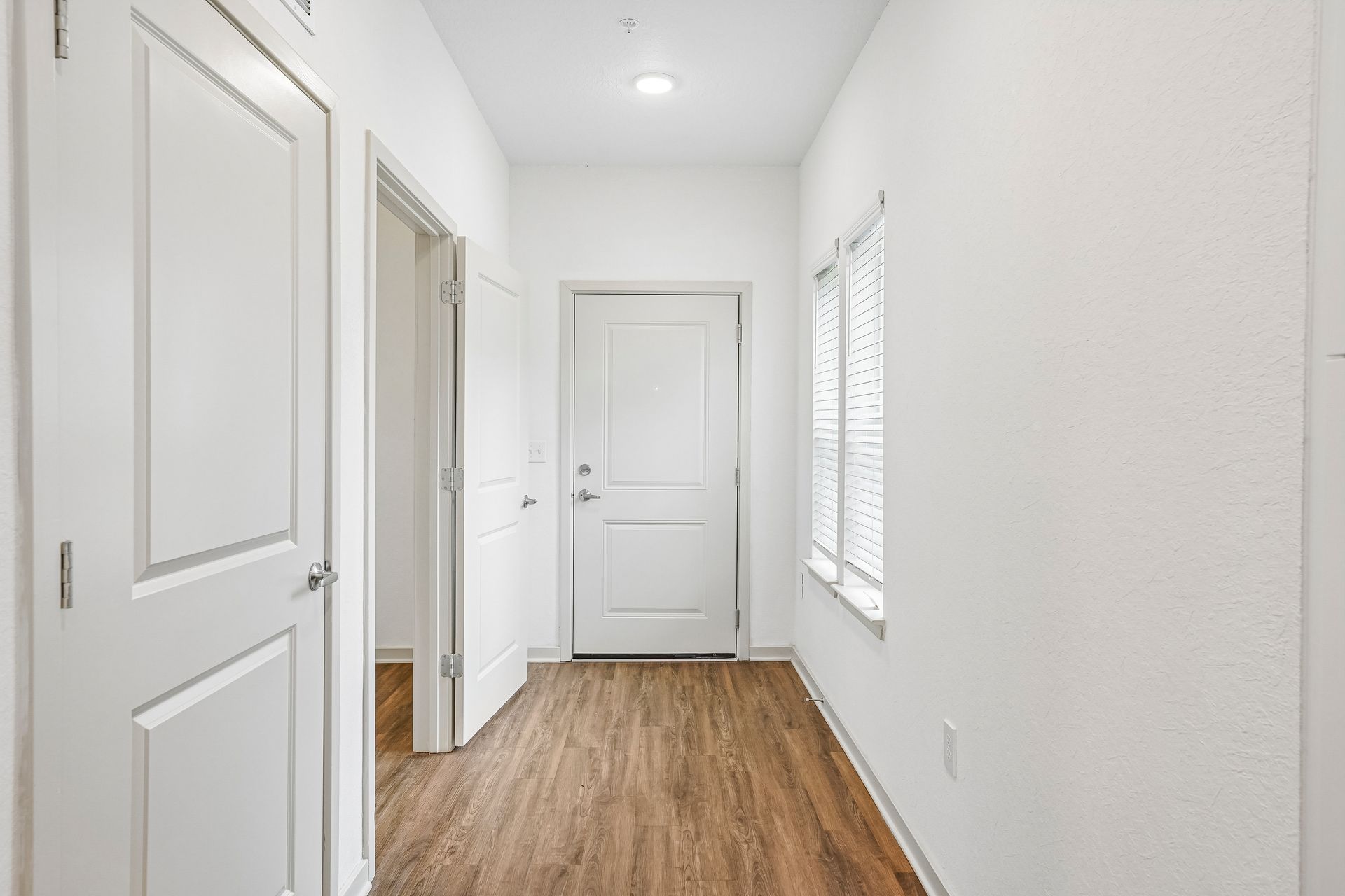 Bright white hallway with wood floors, doors, and a window on the right.