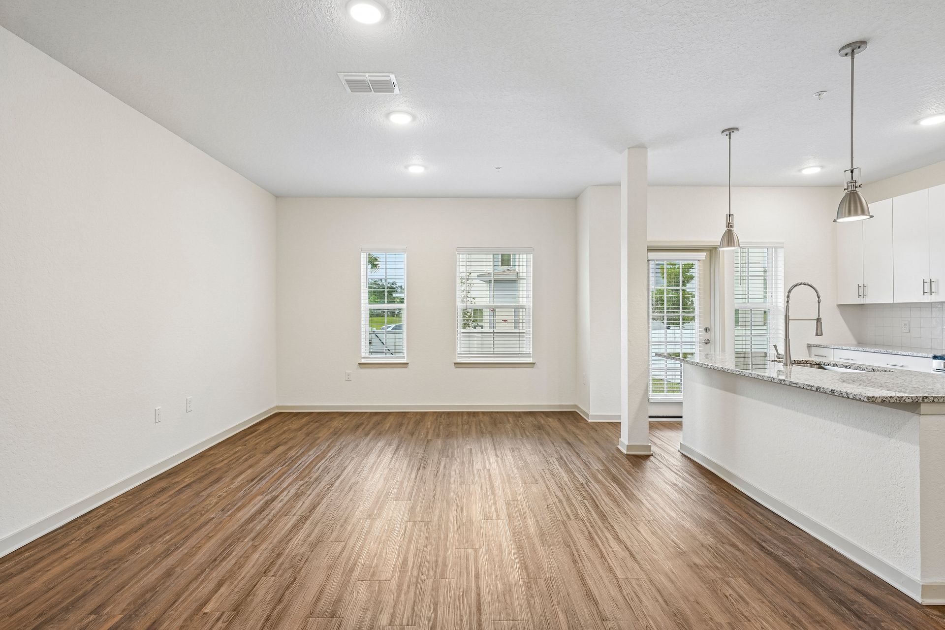 Empty open-concept living area with wood floors, white walls, and a kitchen with a granite island.