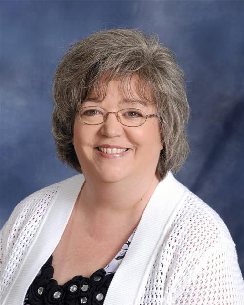 Woman with glasses smiles, wearing a white cardigan over a black and white patterned top, on a blue background.
