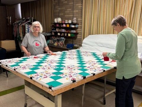 Two women working on a quilt; one at the table, the other standing. Colorful squares on a large table in a room.