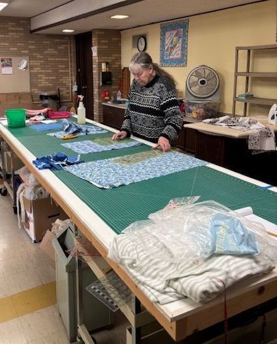 Woman quilting at a large table. She is wearing a sweater, in a well-lit room with fabric and supplies.