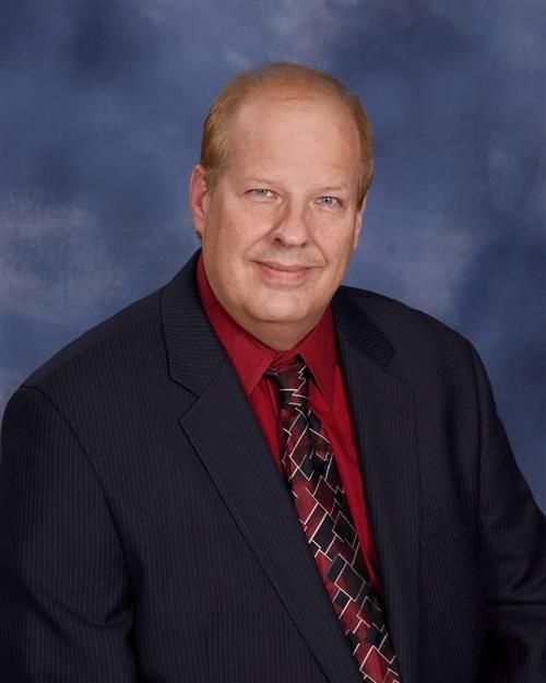 Man in suit smiling at the camera, red shirt and patterned tie, blue background.