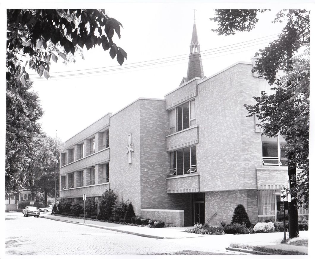Brick building with a spire, St. John new school, on a tree-lined street; a car is parked on the road.