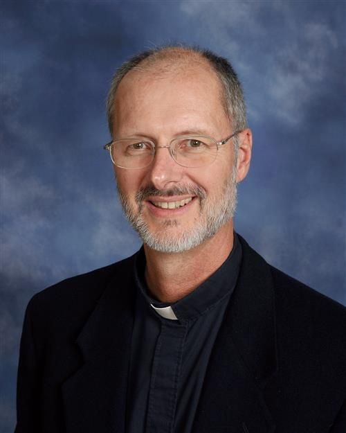 Man with glasses and beard, wearing clerical collar, smiling. Blue background.