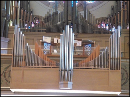 Pipe organ in a church with silver and gold pipes, wooden structure, and stained glass windows.