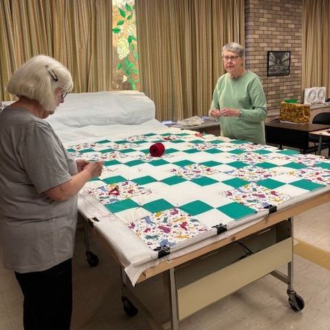 Two women quilting a green and white checkered quilt on a large table in a room.
