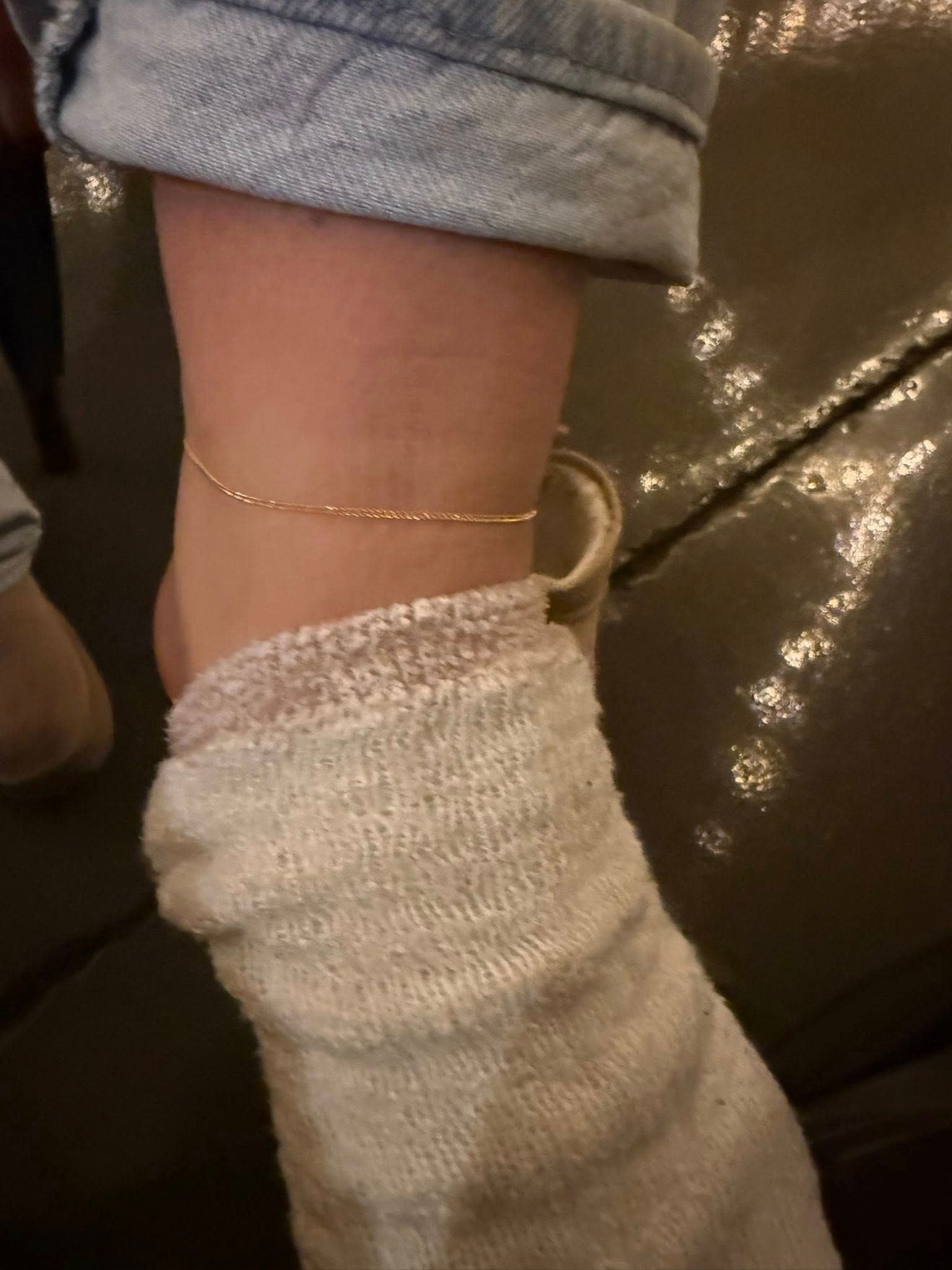 A person’s ankle in a white sock and rolled-up jeans, wearing a thin gold anklet on a dark leather couch.