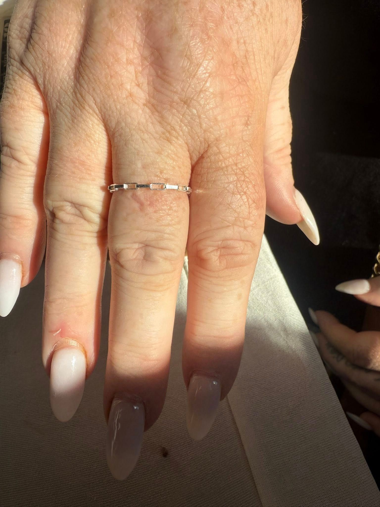 Hand with long white acrylic nails wearing a thin silver ring, resting in sunlight on fabric.