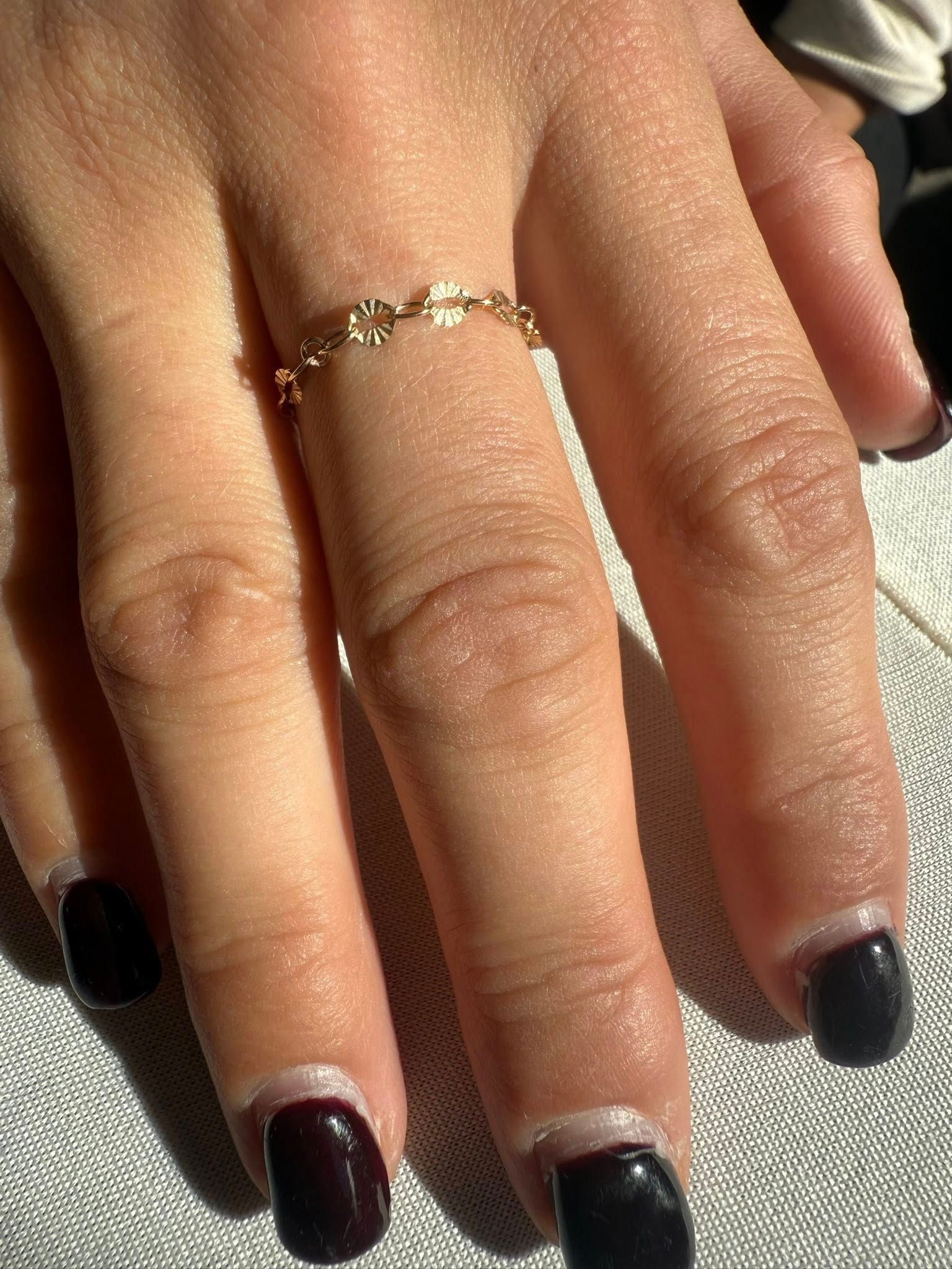 Hand with dark polished nails wearing a delicate gold ring on a sunlit fabric background