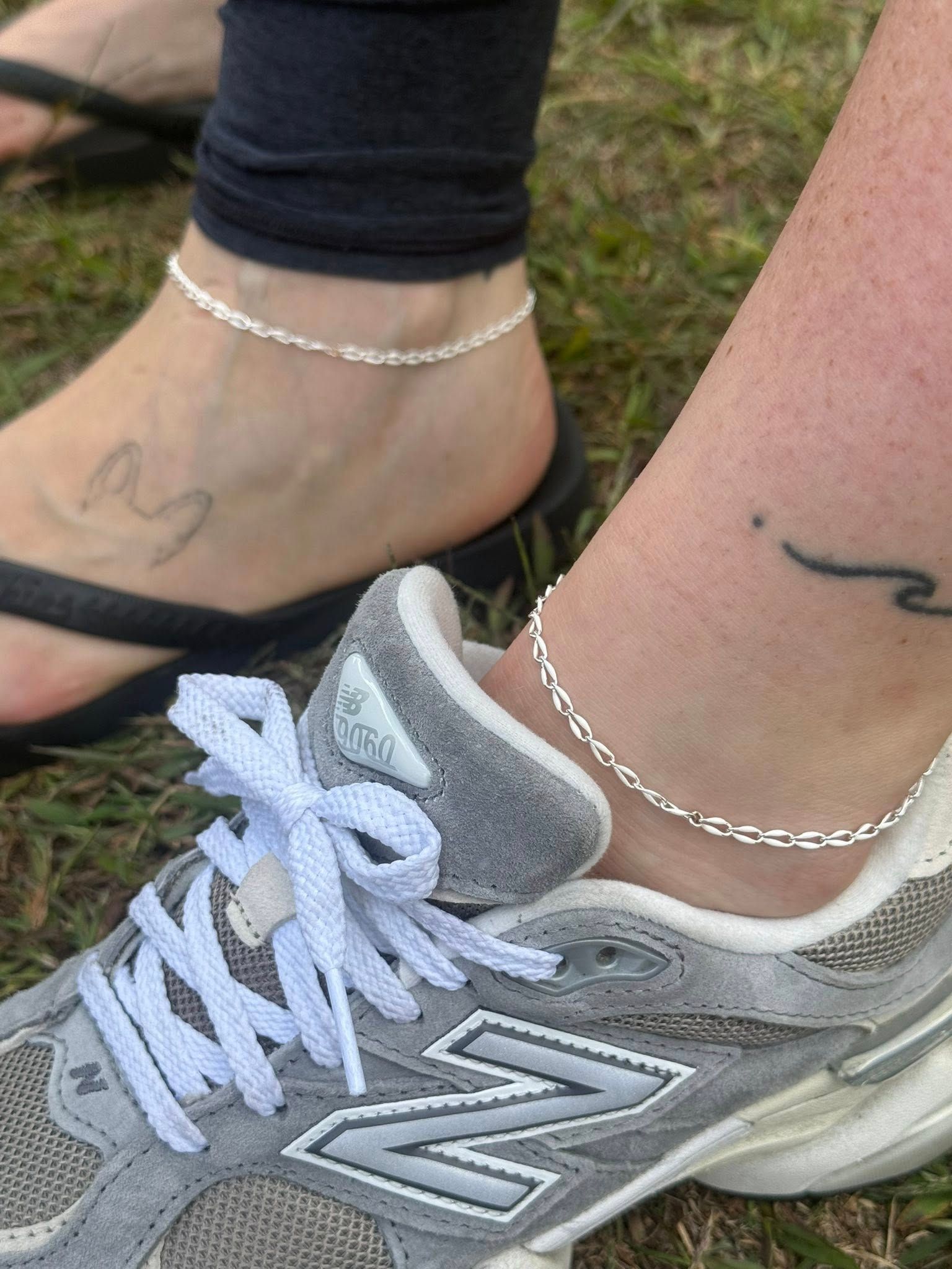 Two ankles with silver anklets above gray New Balance sneakers on grass
