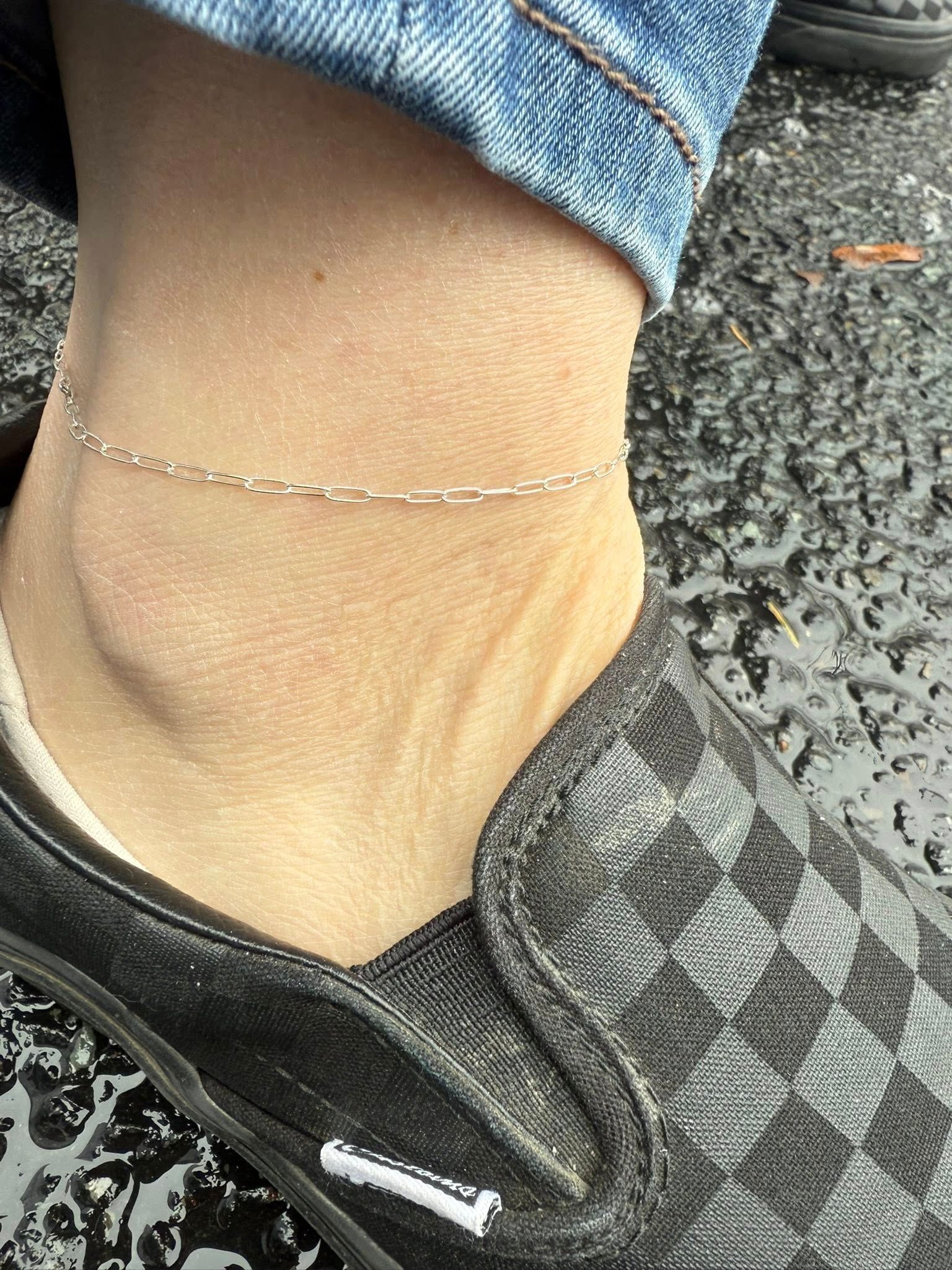 Close-up of an ankle wearing a thin silver anklet and black shoe on gravel