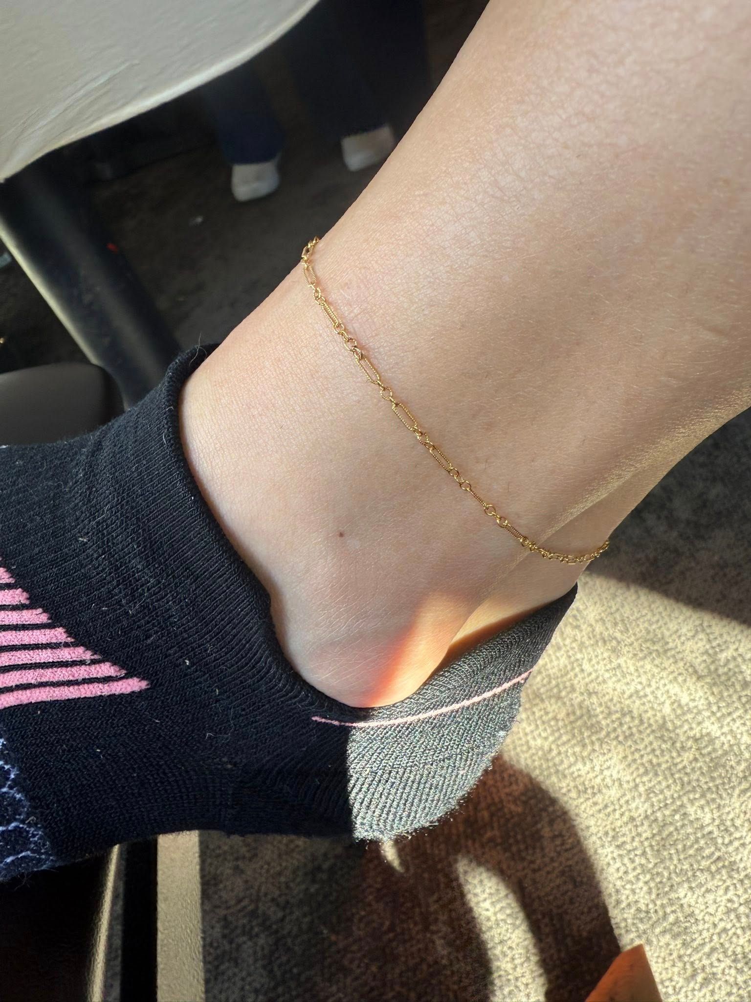 Close-up of a bare ankle wearing a delicate gold anklet and black sock with pink stripes.