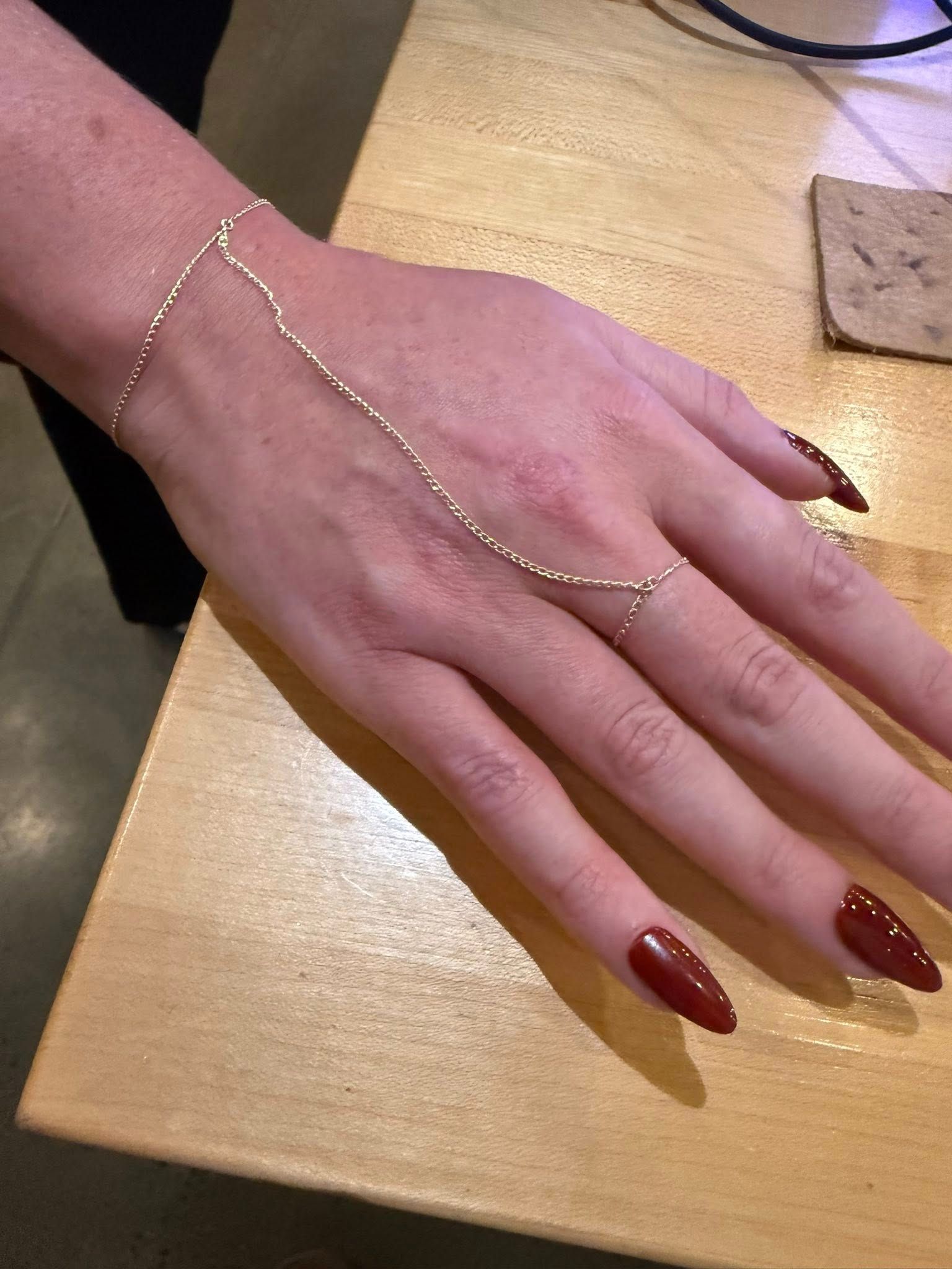 Hand with dark red nails wearing a delicate gold hand chain bracelet on a wooden table