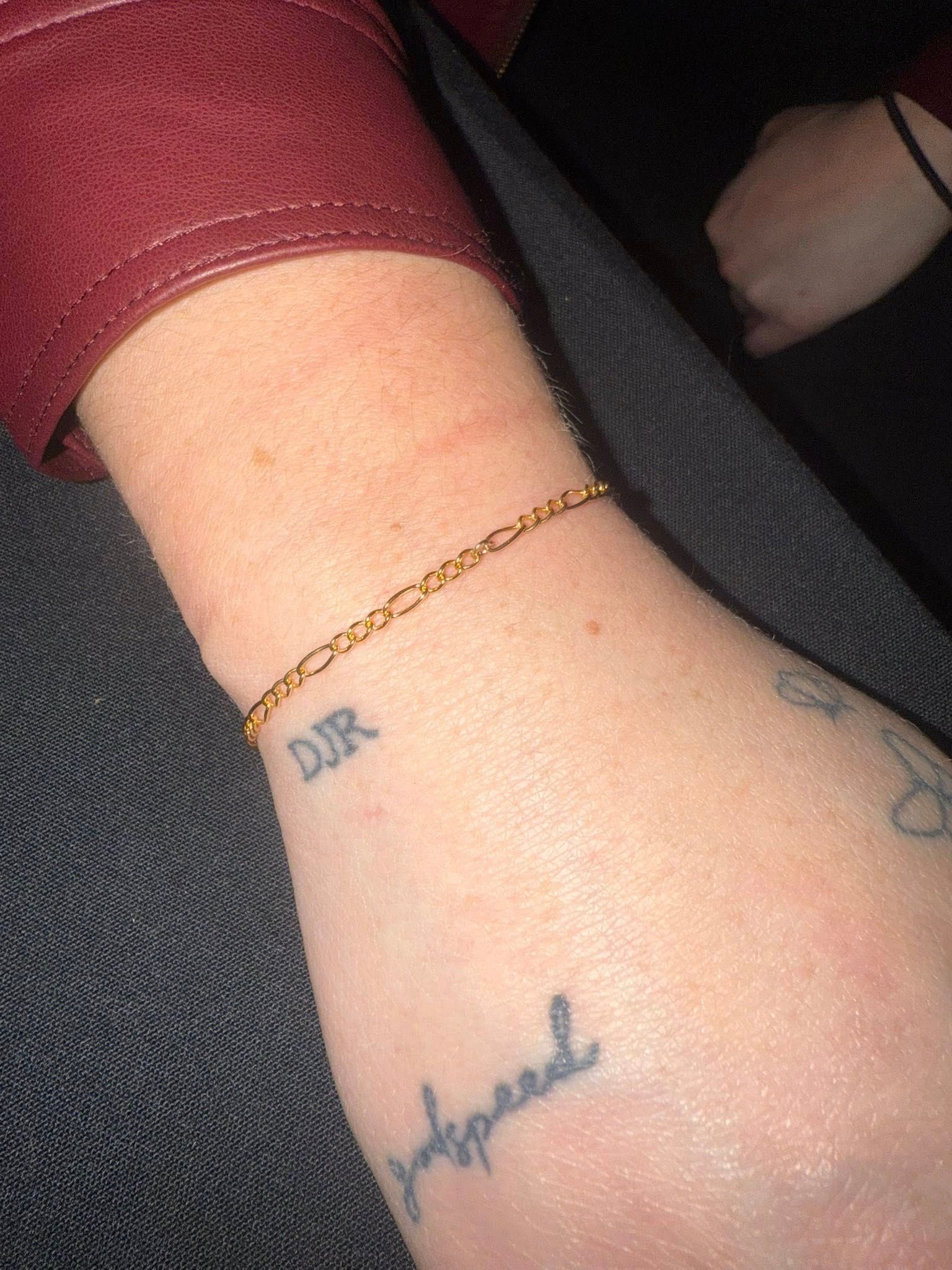 Close-up of a wrist with a thin gold bracelet and small tattoos against a dark seat.