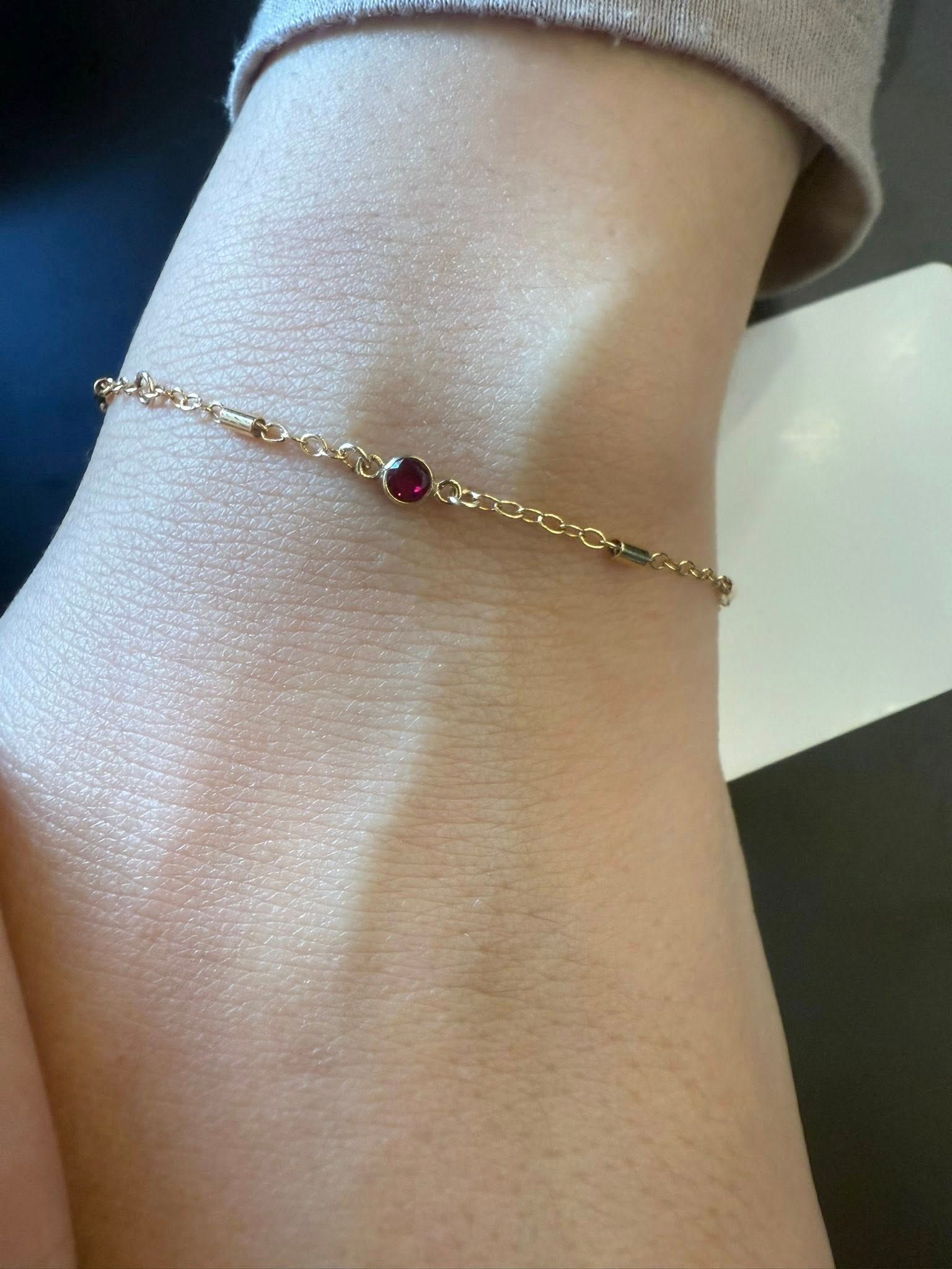 Gold bracelet with a small red gemstone on a wrist with a beige sleeve.