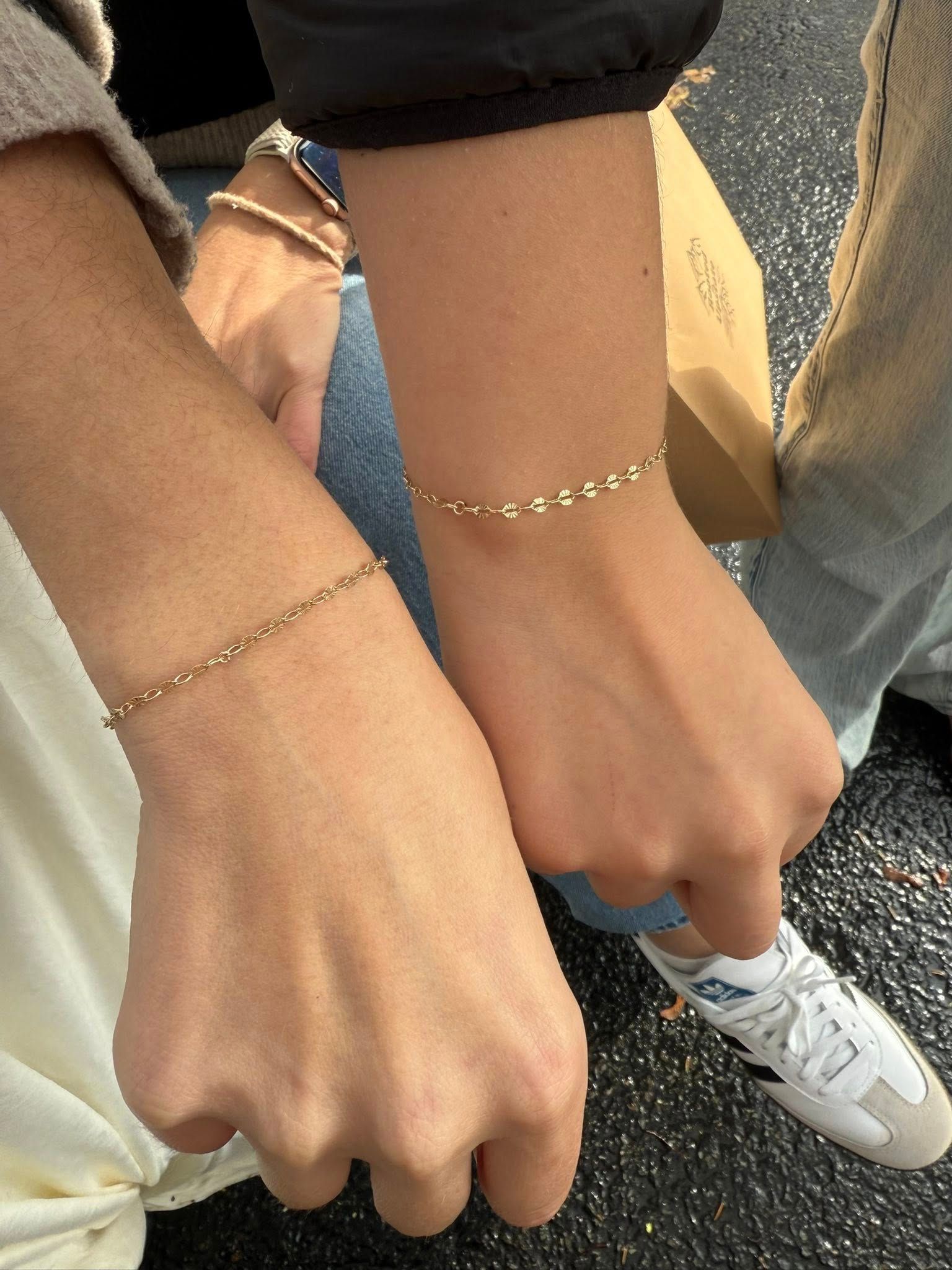 Two wrists with delicate bracelets held together over a street, beside a white sneaker and jeans