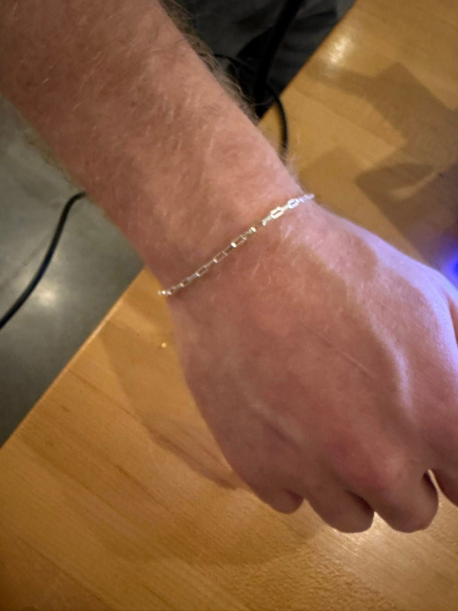 Wrist wearing a thin silver chain bracelet over a wooden table.