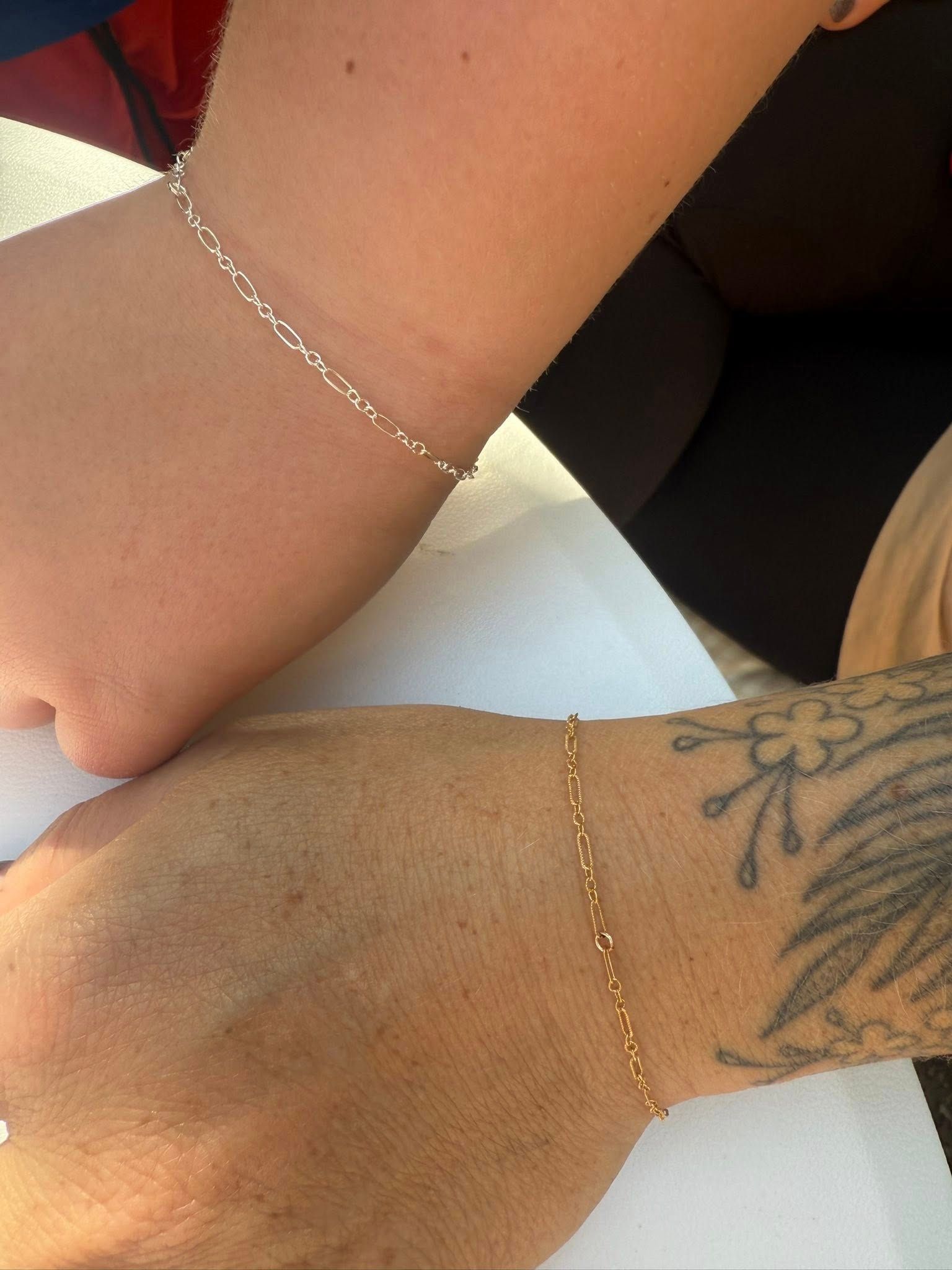Two arms touching wrists, one with a silver bracelet and one with a gold chain and wrist tattoo.
