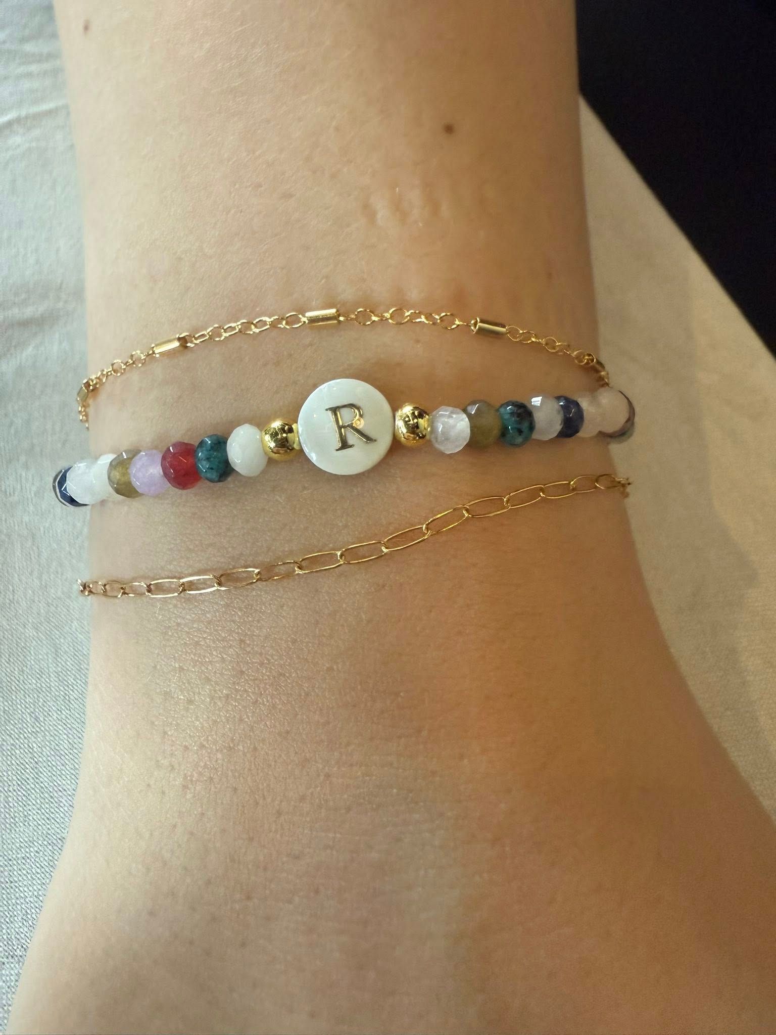 Beaded bracelet with gold chains on a wrist, featuring a white center charm with a gold letter R.