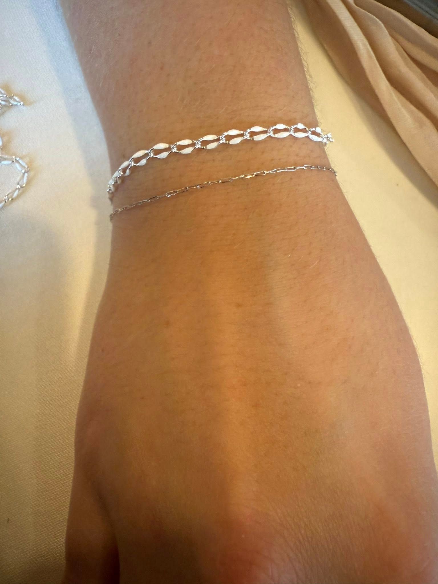 Close-up of a wrist wearing two silver chain bracelets on a beige fabric background