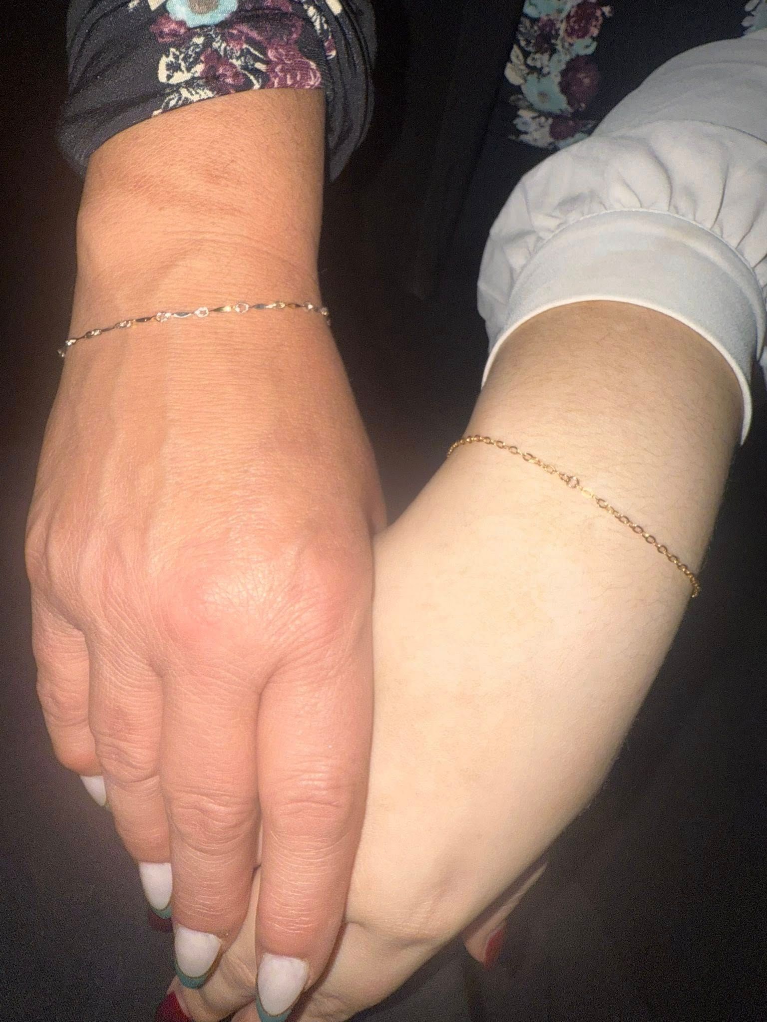 Two hands with matching bracelets clasped together, one in a floral sleeve and one in a white sleeve.